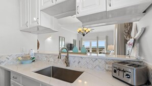 A modern kitchen with white cabinetry, a herringbone tile backsplash, a stainless steel sink, a toaster, and a view of the ocean through large windows.