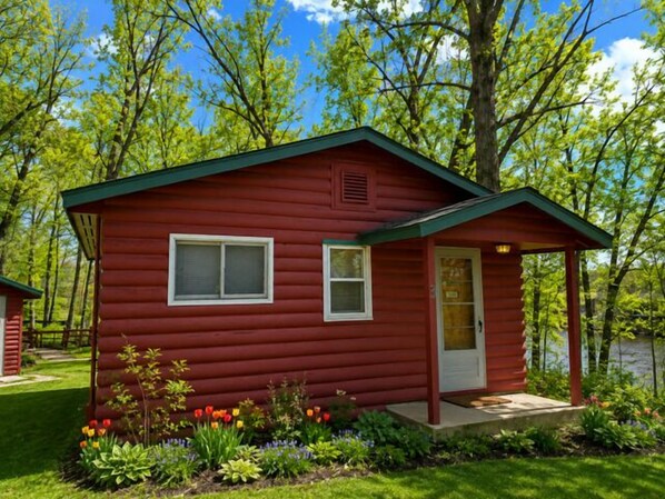 A small, red log cabin with a porch, a door, and windows.