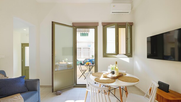 A modern living room with a sofa, a dining table with chairs, a TV mounted on the wall, and a sliding glass door leading to an outdoor area. - Elartis Apartment (Chania)