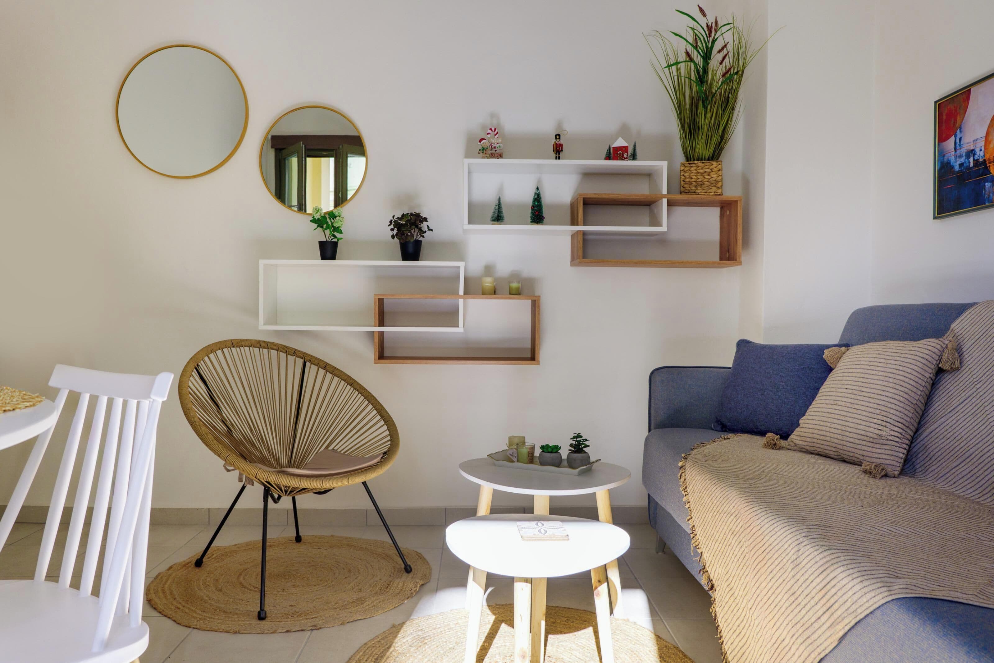 Ein modernes Wohnzimmer mit einem weißen Sofa, einem runden Spiegel und einem Rattanstuhl. Regale mit Pflanzen und Dekorationselementen sowie ein kleiner weißer Tisch mit einem Buch.