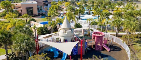 Un parque infantil con toboganes y estructuras para escalar, rodeado de palmeras y una cerca blanca.