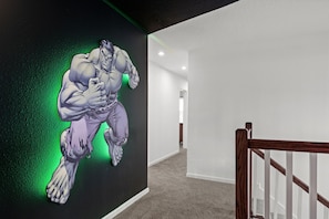 A hallway with a large wall mural of a muscular figure, a wooden staircase, and a carpeted floor.