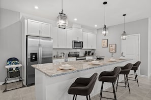 A modern kitchen with white cabinets, a stainless steel refrigerator, a microwave, and a granite countertop. There are three bar stools and a high chair.