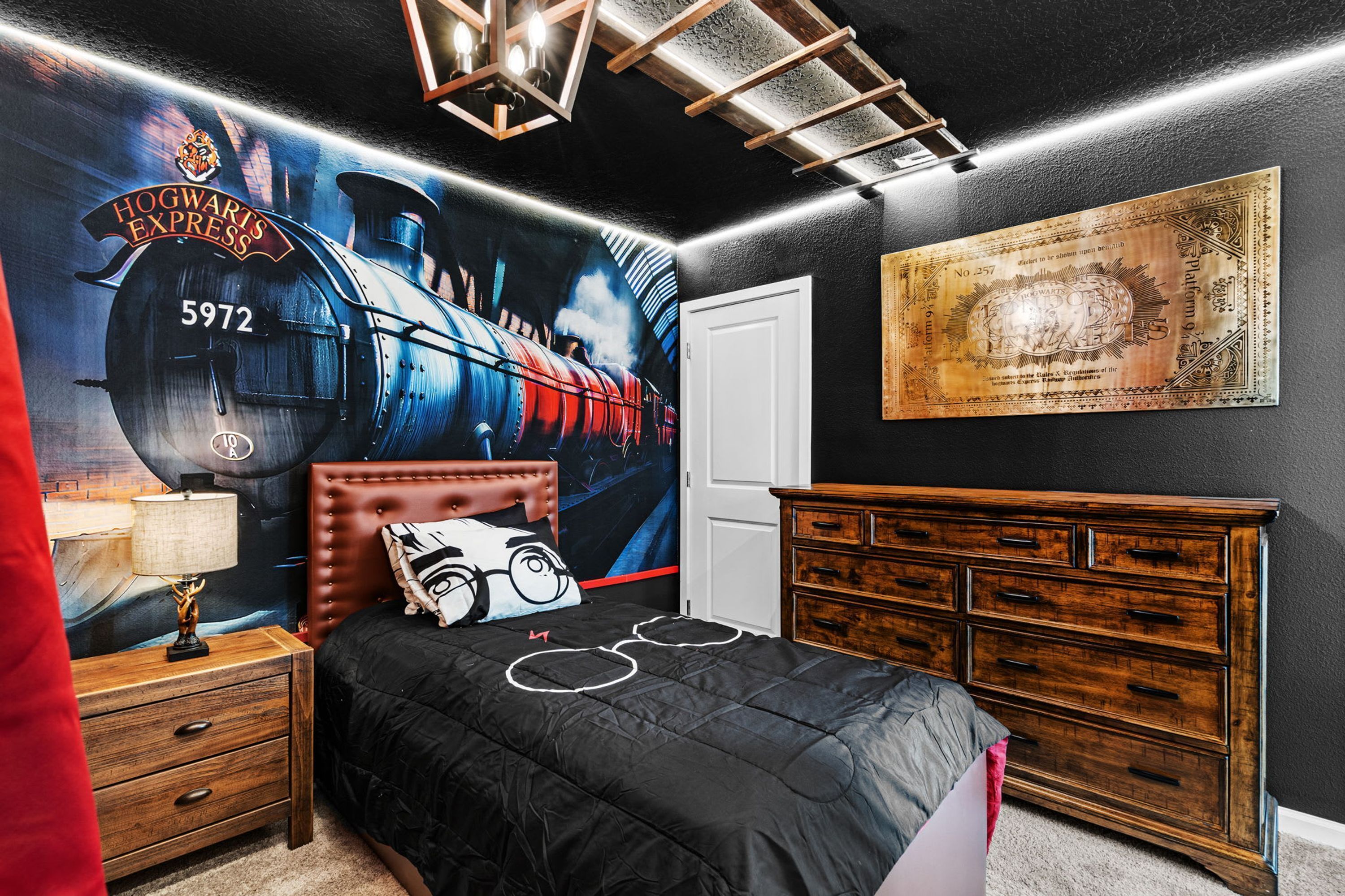 A bedroom with a Hogwarts Express mural, a wooden dresser, a bed with a black and white themed duvet, and a red curtain.