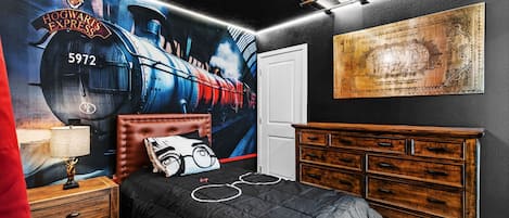 Une chambre avec une fresque représentant le Poudlard Express, une commode en bois, un lit avec une couette aux motifs noir et blanc, et un rideau rouge.