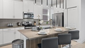 Cozinha moderna com armários brancos, eletrodomésticos de aço inoxidável, ilha central com banquetas e iluminação pendente.