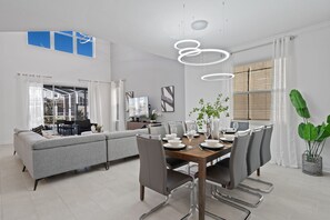 A modern dining area with a wooden table set for six, surrounded by grey upholstered chairs. A grey sofa is positioned in the background, and a large window with sheer curtains is visible. - Chic 5bed Retreat Perfect for Family Getaways (Kissimmee)
