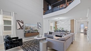 Uma sala de estar espaçosa com um sofá cinzento, uma poltrona preta e uma mesa de centro. Há uma escadaria que leva a um andar superior com uma varanda.