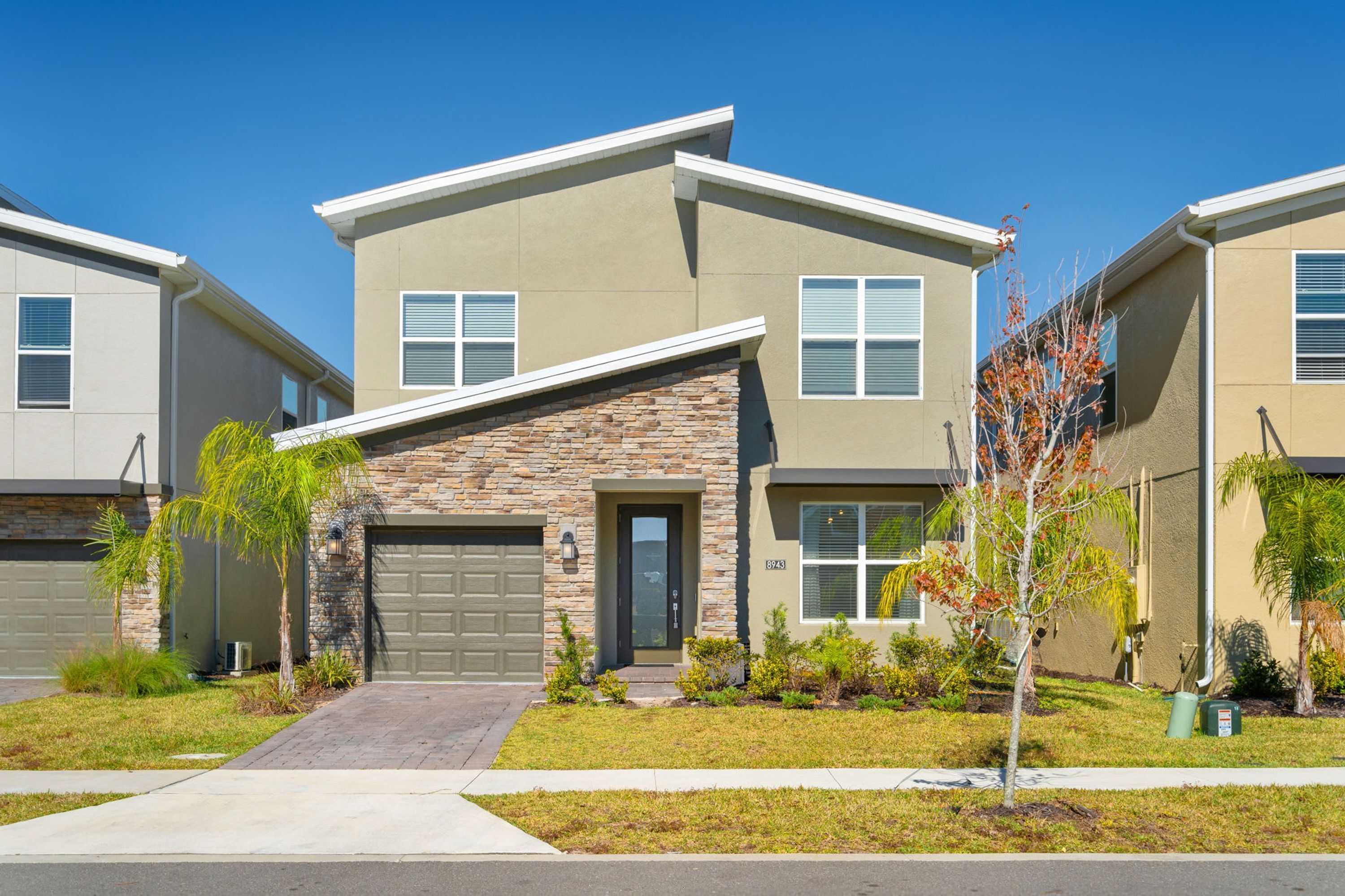 A modern two-story residential building with a stone facade, a garage, and a well-maintained lawn with small trees and shrubs.