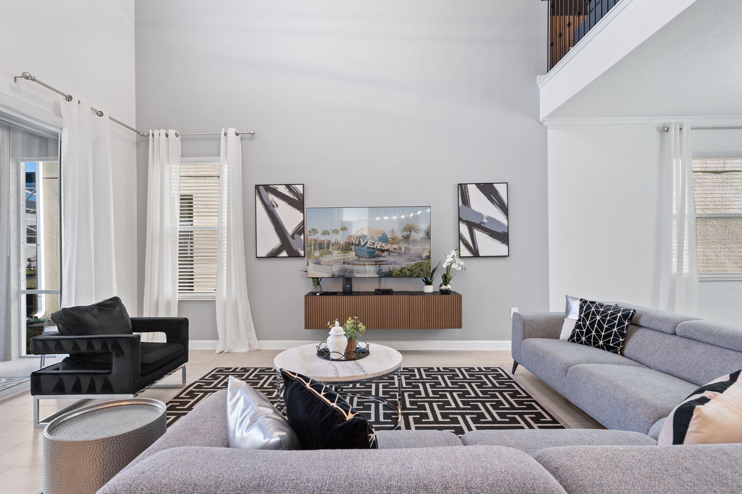 A modern living room with a grey sofa, a black armchair, and a coffee table. There is a large TV mounted on the wall, and a window with white curtains.