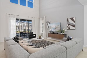 A spacious living room with a grey sofa, a coffee table, and a TV on a wooden stand. There are two large windows with curtains, and a painting on the wall. - Chic 5bed Retreat Perfect for Family Getaways (Kissimmee)