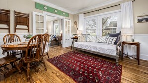 A room with a wooden dining table and chairs, a bed with a red patterned rug, and a window with white curtains.
