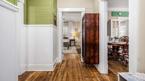 A hallway with wooden flooring, white walls, and a green accent wall. There is a wooden door on the right and a dining area with a table and chairs visible in the background.