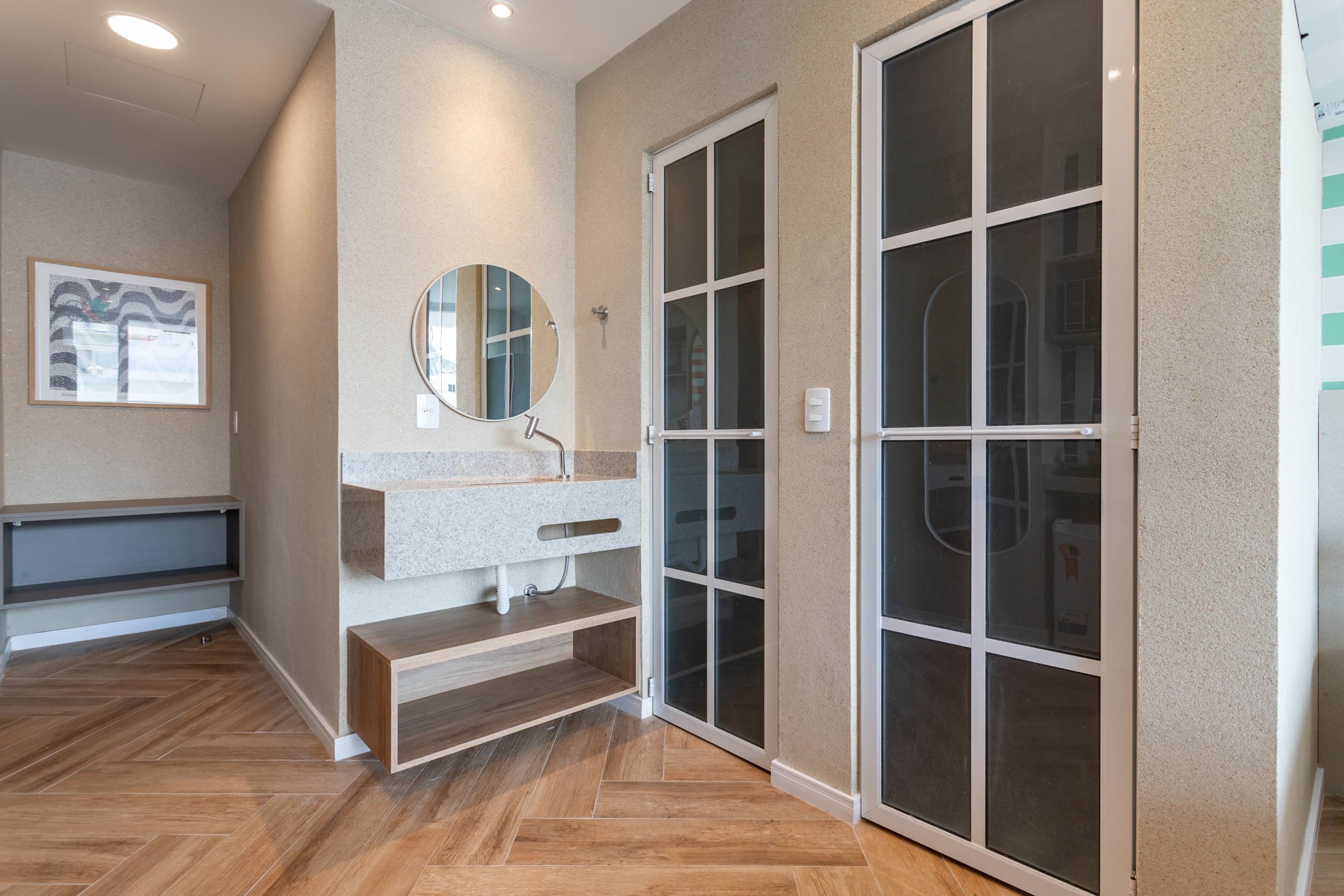 Een moderne badkamer met een houten visgraatvloer, een granieten aanrechtblad, een ronde spiegel en een glazen schuifdeur naar een berging.
