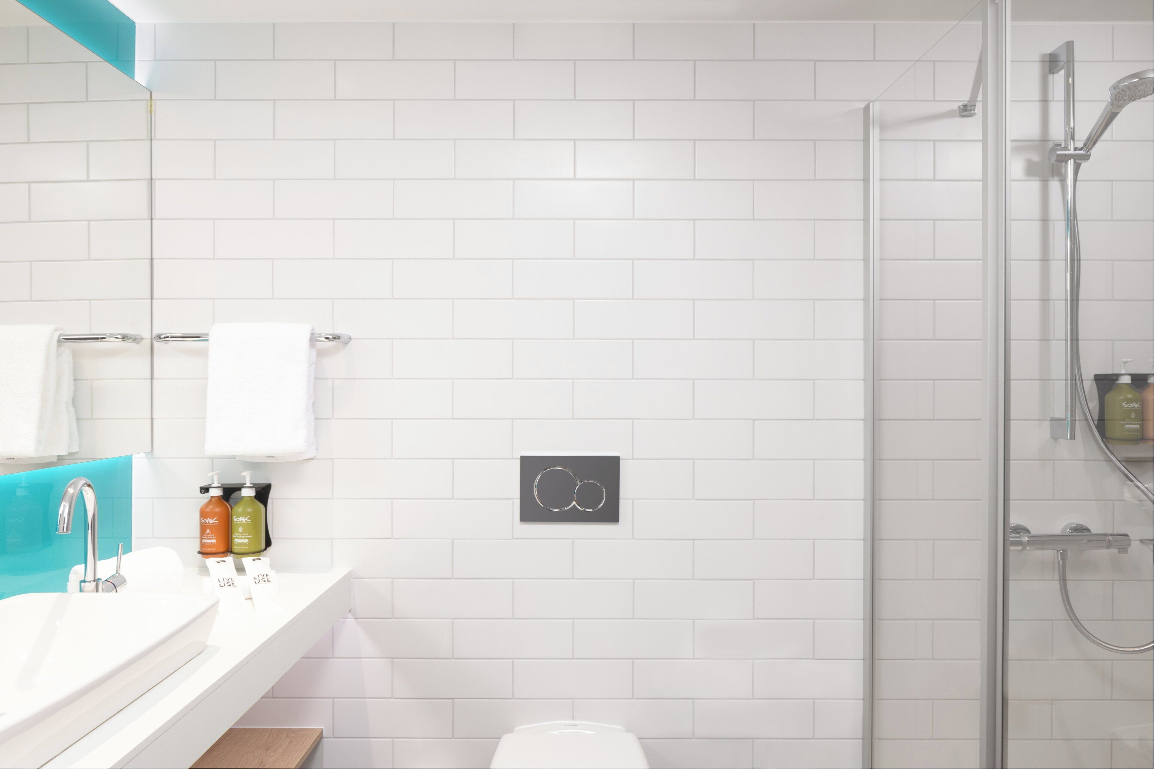 A modern bathroom with a white tiled wall, a glass shower enclosure, a white sink with a chrome faucet, and a mirror above the sink.