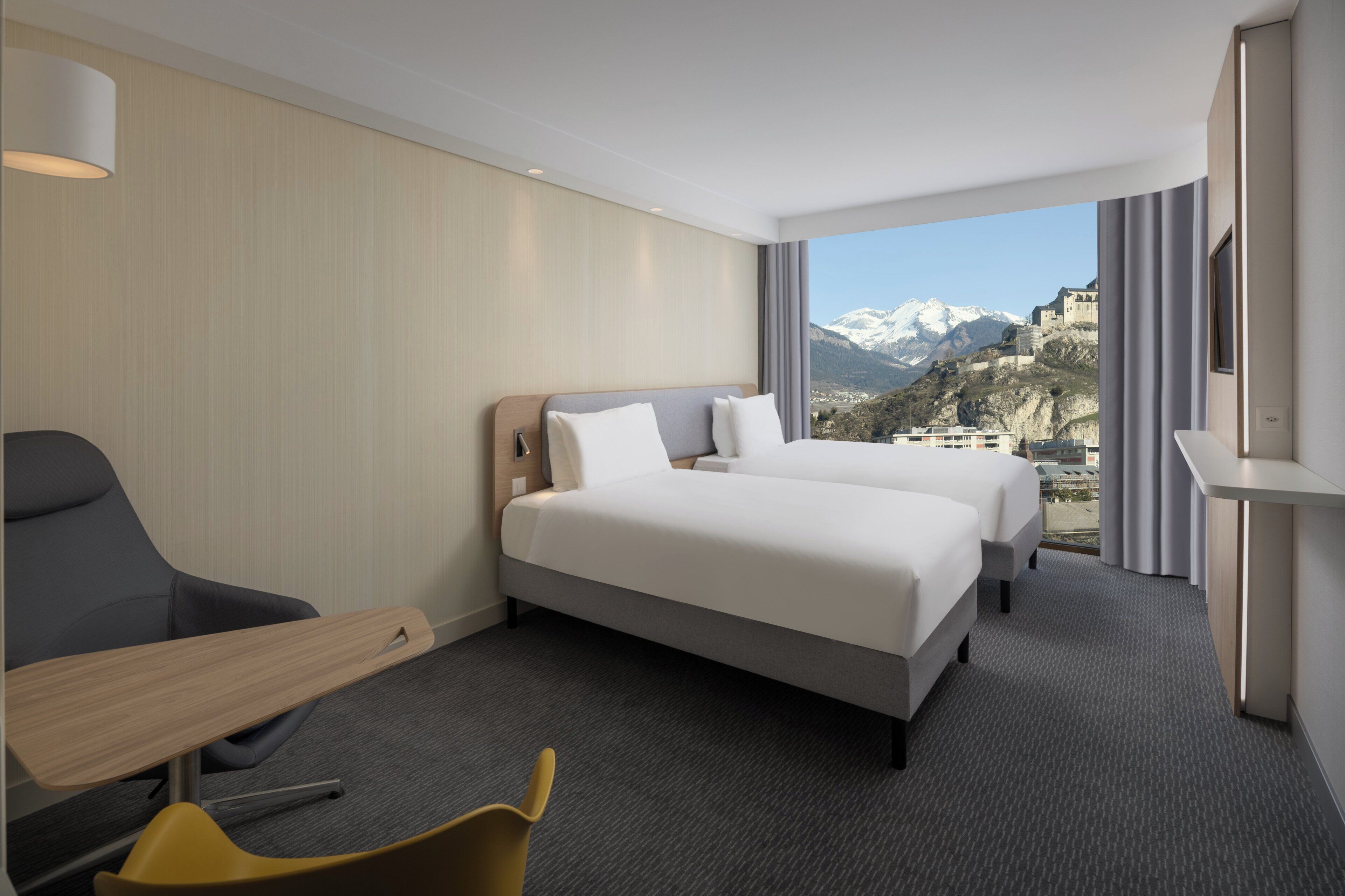 Habitación de hotel con dos camas, un escritorio, una silla y un amplio ventanal con vistas a la montaña.