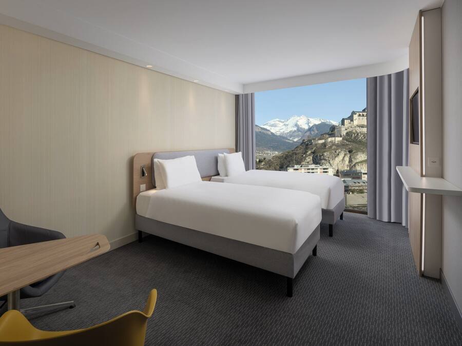 Hotelkamer met twee bedden, een bureau, een stoel en een groot raam met uitzicht op de bergen.