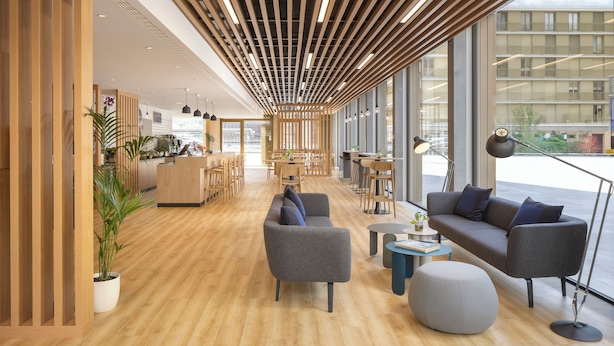 Ein modernes Interieur mit Holzboden, grauen Sofas, einem runden Hocker und großen Fenstern mit Blick auf die Stadt.