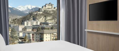 A hotel room with a large bed, a TV mounted on the wall, and a view of a mountain and buildings.