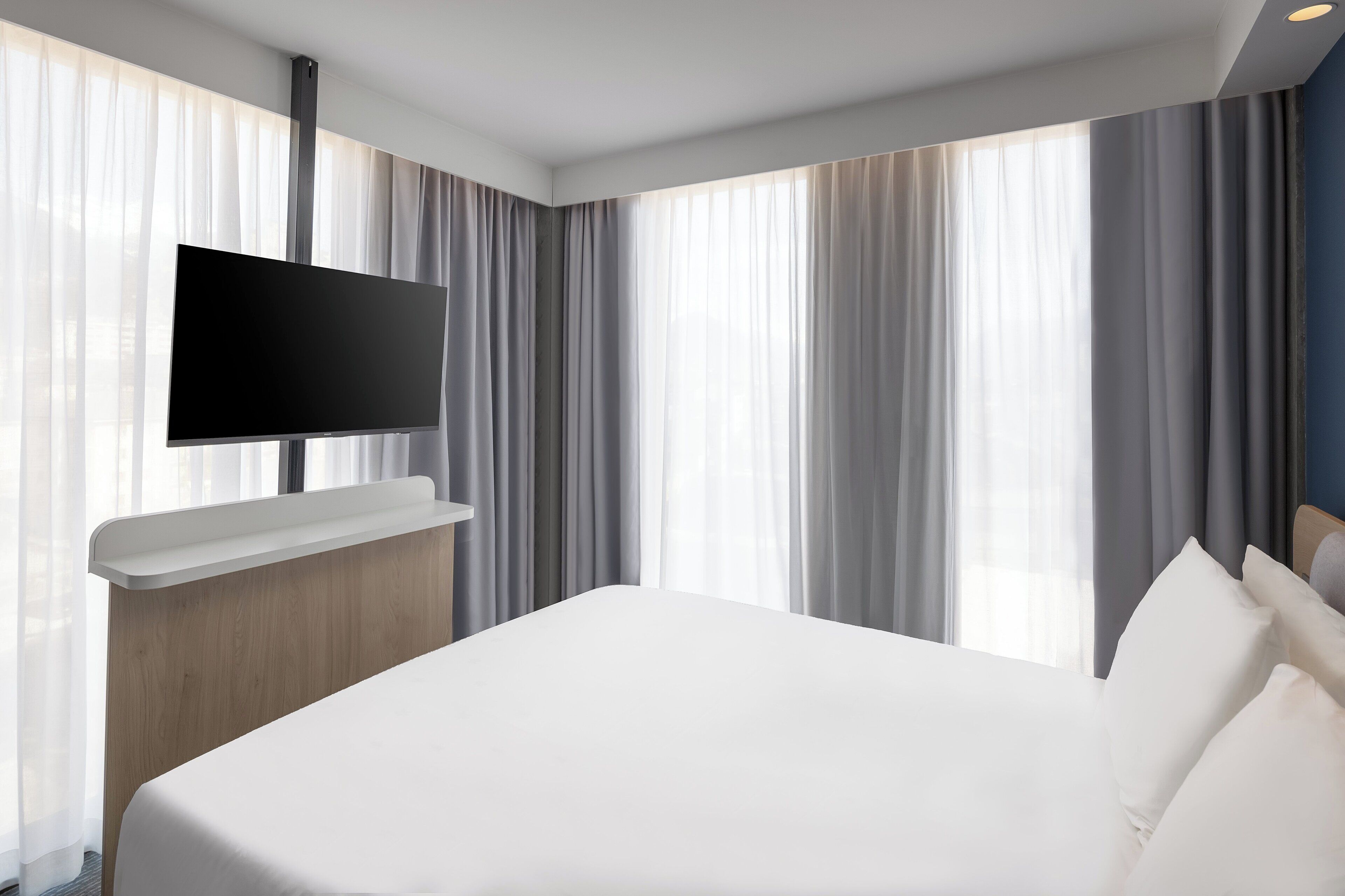 A modern hotel room with a large bed, a flat-screen TV mounted on a wooden console, and large windows with sheer curtains.