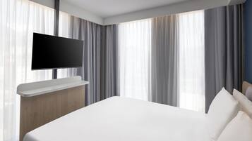 A modern hotel room with a large bed, a flat-screen TV mounted on a wooden console, and large windows with sheer curtains.