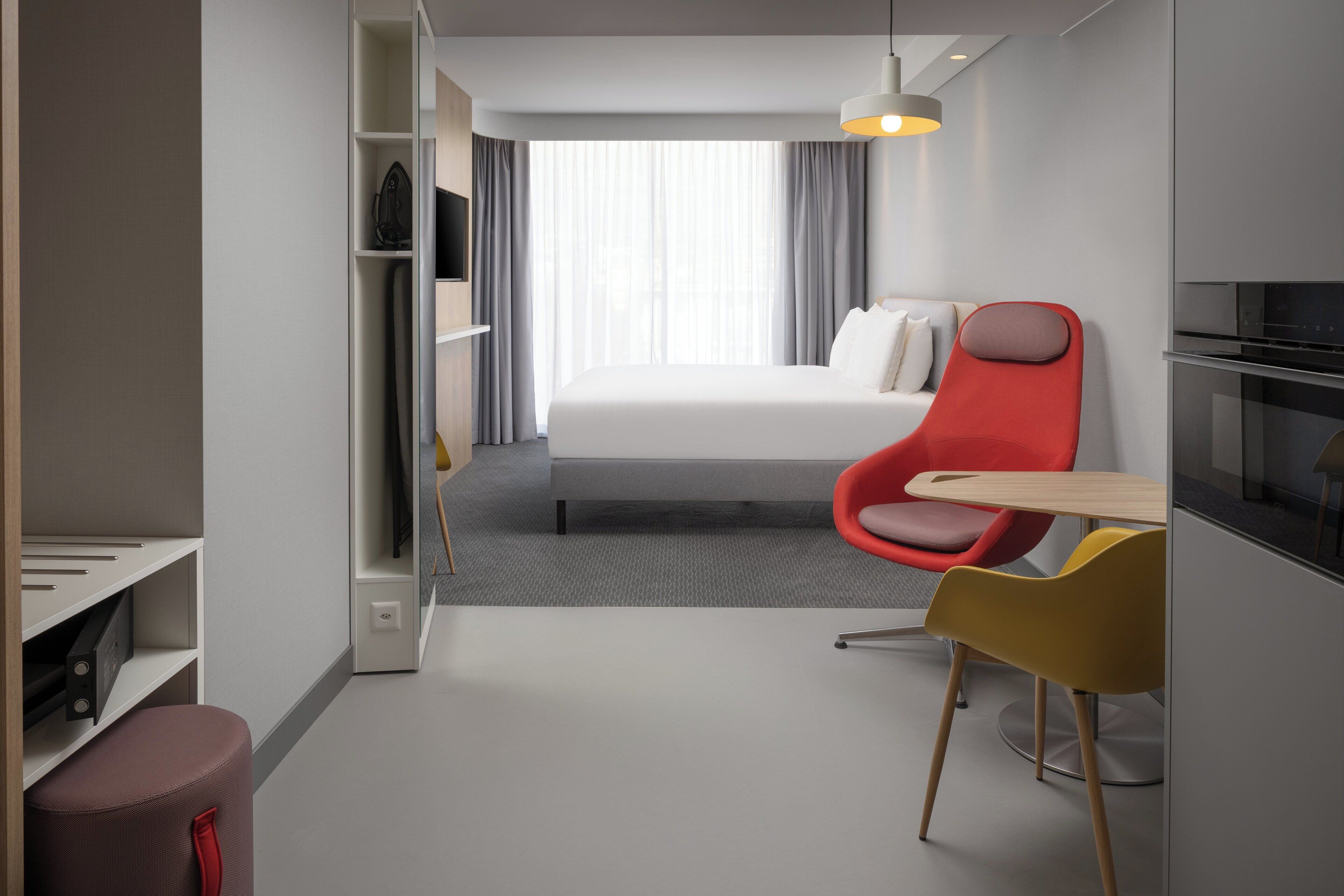 Une chambre d’hôtel moderne avec un lit, une chaise rouge, une chaise jaune, une petite table et un téléviseur.