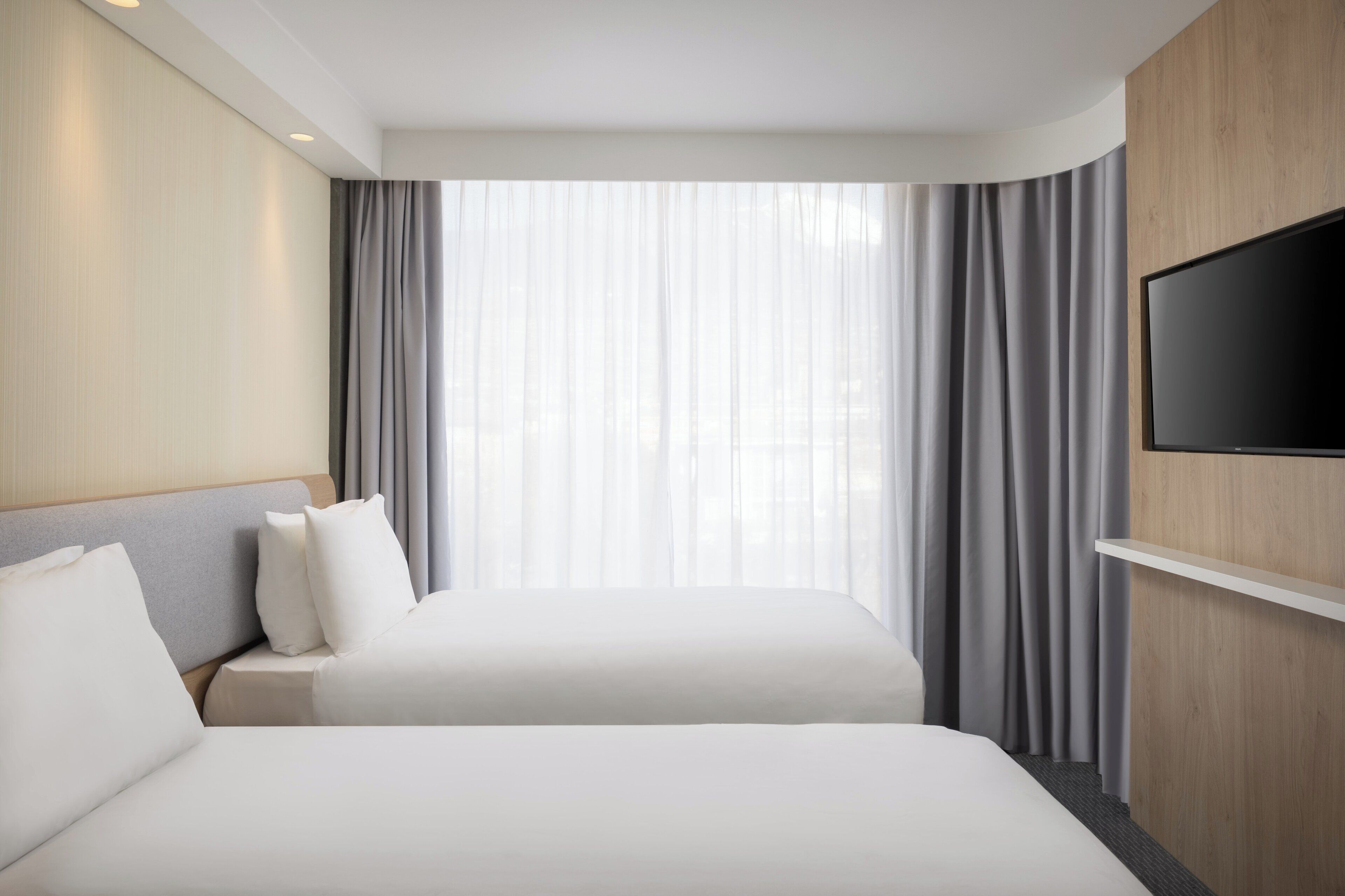 Une chambre d’hôtel avec deux lits, un téléviseur à écran plat fixé au mur et une grande fenêtre avec des rideaux légers.