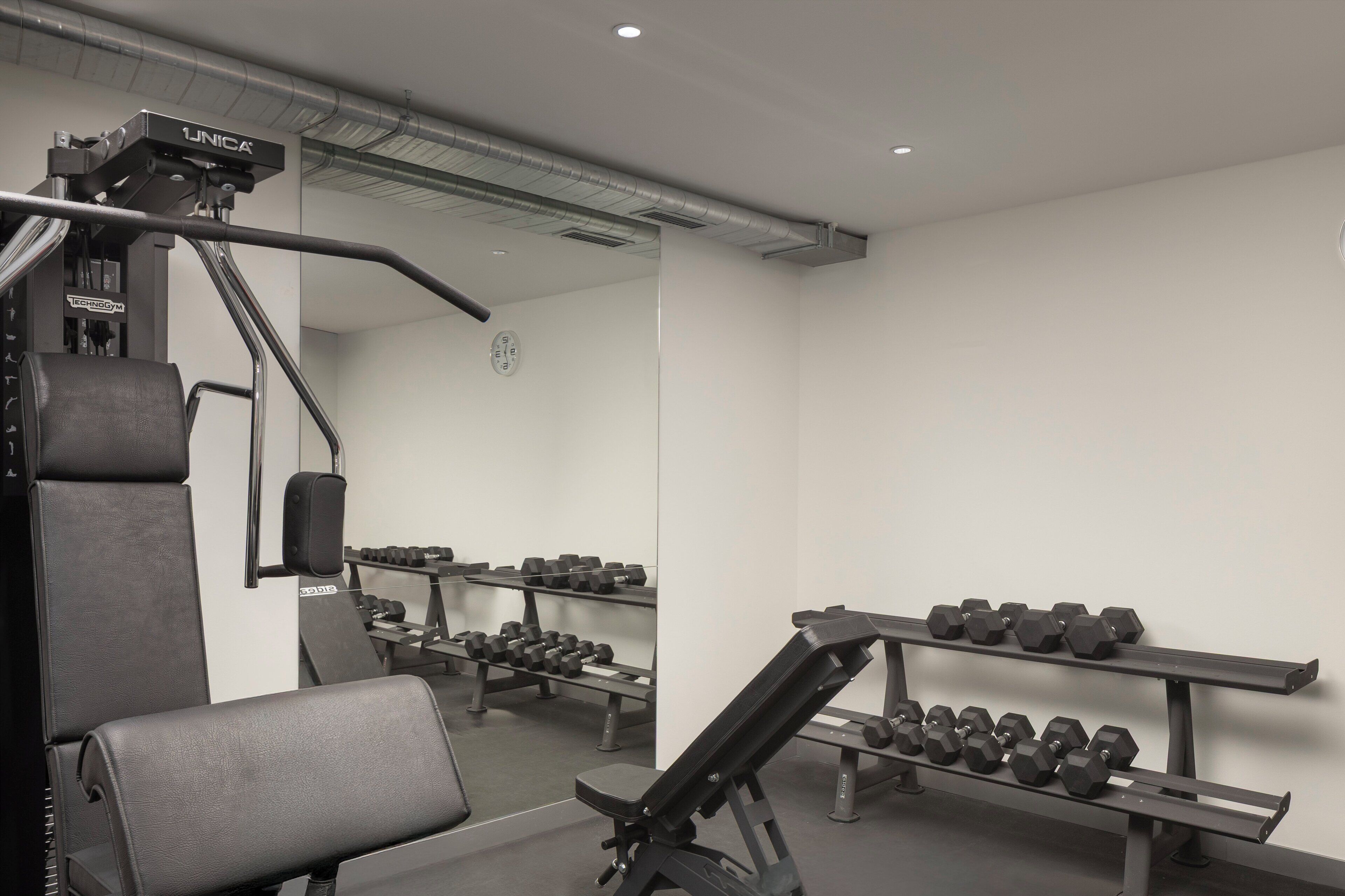 Un gimnasio con equipo de levantamiento de pesas, bancos con mancuernas y un espejo en la pared.