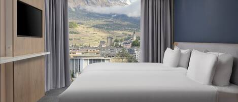 A modern hotel room with a large bed, a flat-screen TV mounted on the wall, and a view of a mountainous landscape.
