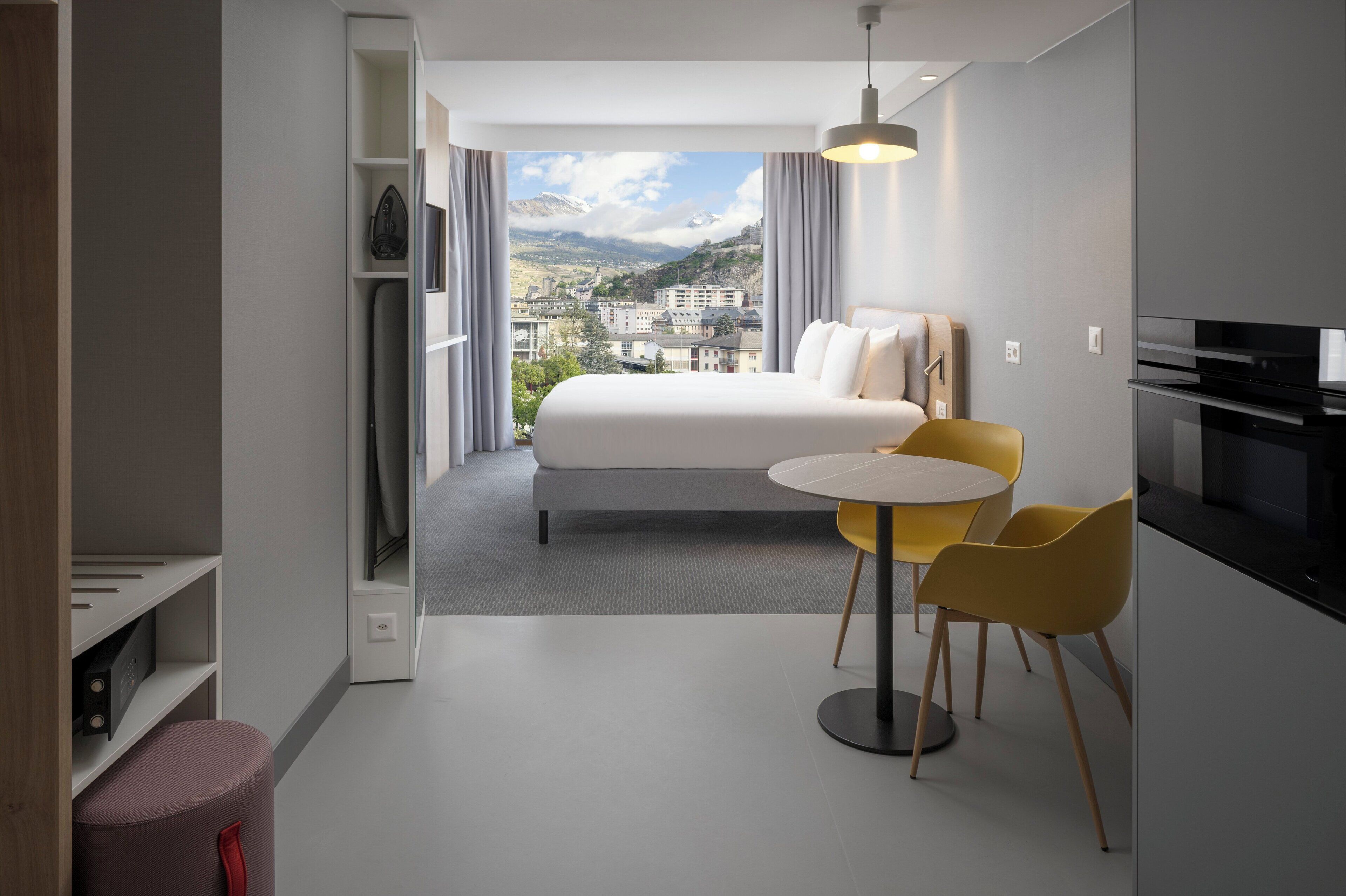 Une chambre d’hôtel moderne avec un lit, une table à manger entourée de chaises jaunes, un coin cuisine et une vue sur le paysage urbain à travers de grandes fenêtres.