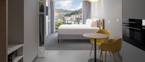 Ein modernes Hotelzimmer mit einem Bett, einem Esstisch mit gelben Stühlen, einer Küchenzeile und Blick auf eine Stadtlandschaft durch große Fenster.