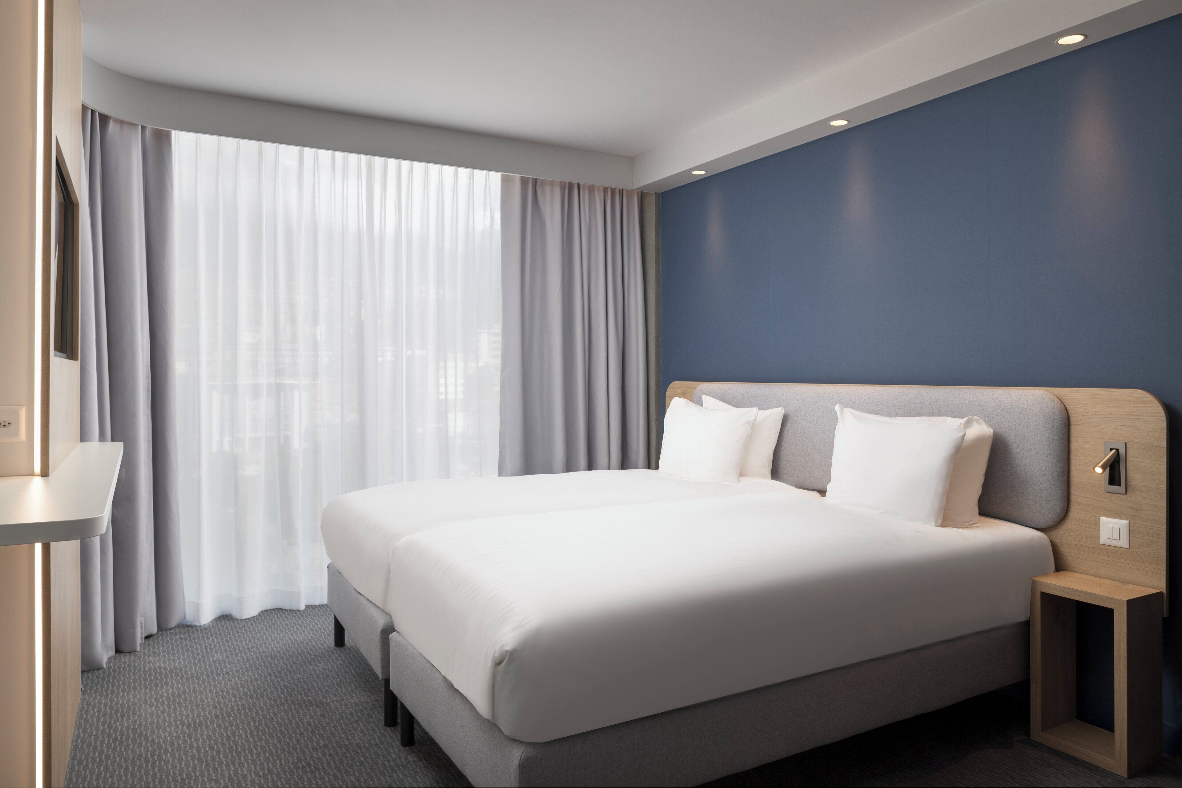 Una habitación de hotel moderna con una cama grande, una mesita de noche, un espejo y un ventanal con cortinas ligeras.