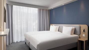 Una habitación de hotel moderna con una cama grande, una mesita de noche, un espejo y un ventanal con cortinas ligeras.