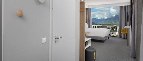 A hotel room with a bed, a small table, a chair, and a view of mountains.