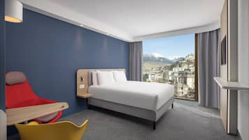 A modern hotel room with a large bed, a desk, a chair, and a view of a cityscape with mountains.