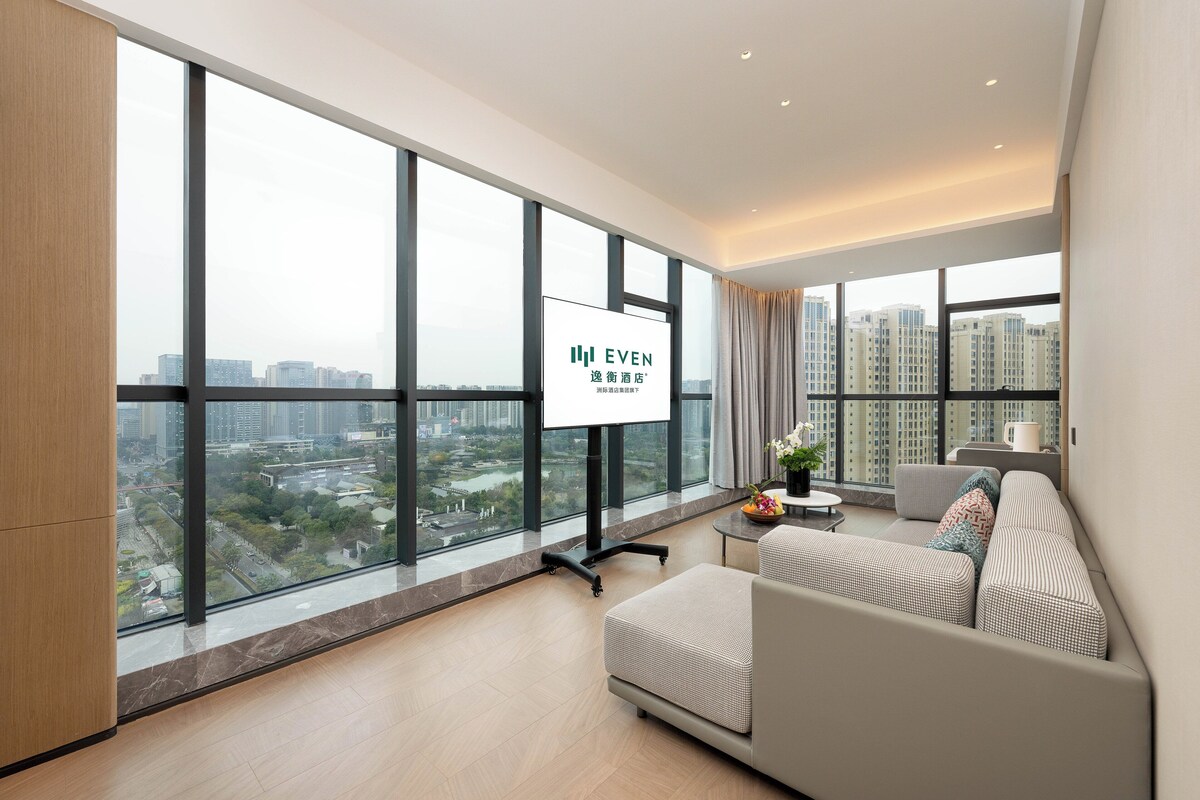 A modern living room with large windows, a sofa, and a TV screen displaying a hotel name.
