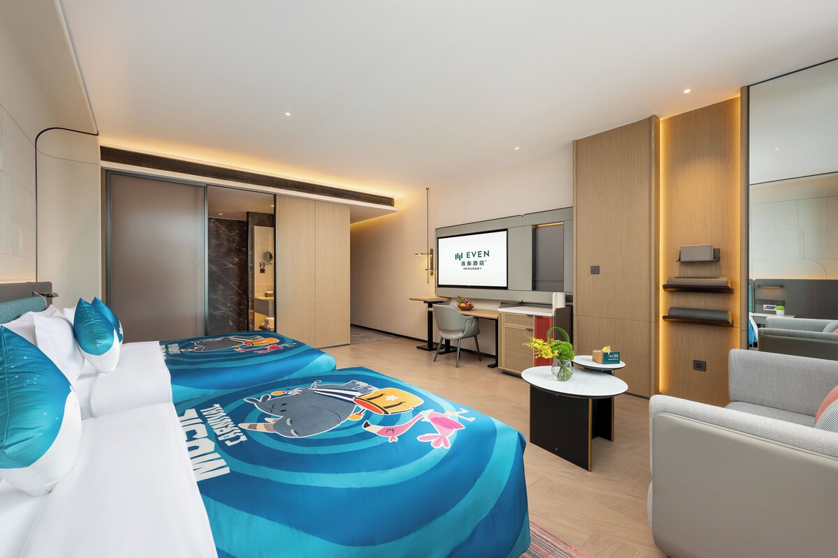 A modern hotel room with two beds, a desk, a TV, and a seating area.