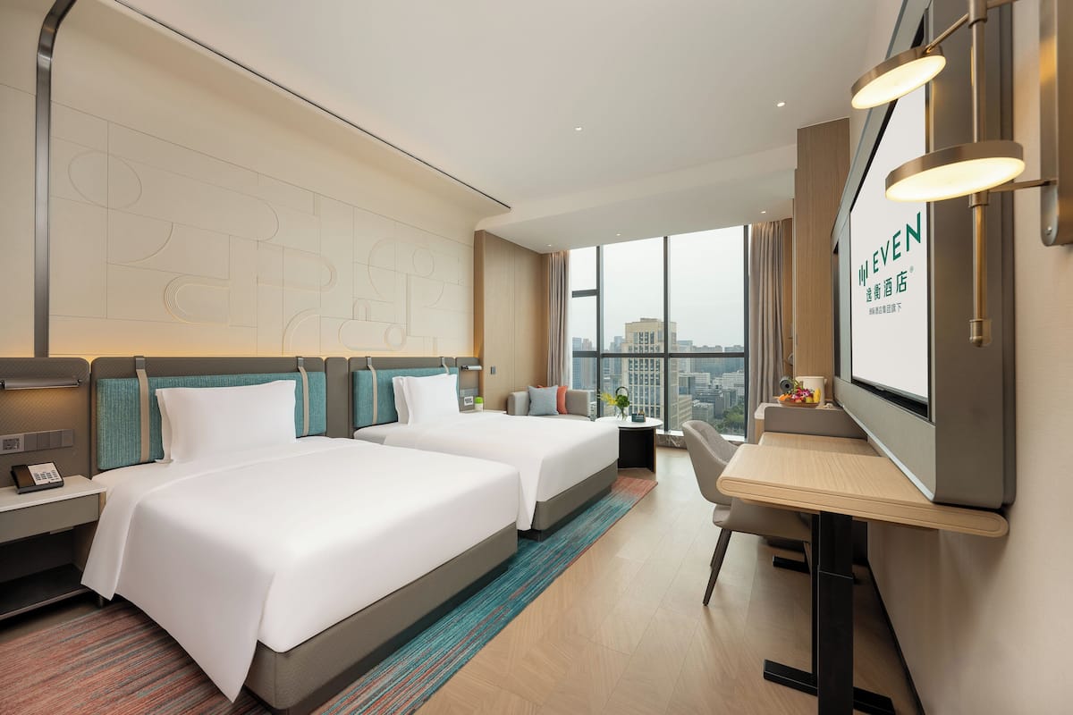 Habitación de hotel con dos camas, un escritorio, una silla, un televisor y un ventanal con vistas a la ciudad.
