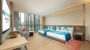 A modern hotel room with two beds, a desk, a chair, a small table with fruit, and a large window offering a city view.