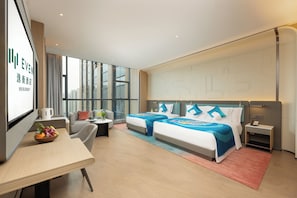Une chambre d’hôtel moderne équipée de deux lits, d’un bureau, d’une chaise, d’une petite table avec des fruits et d’une grande fenêtre donnant sur la ville.