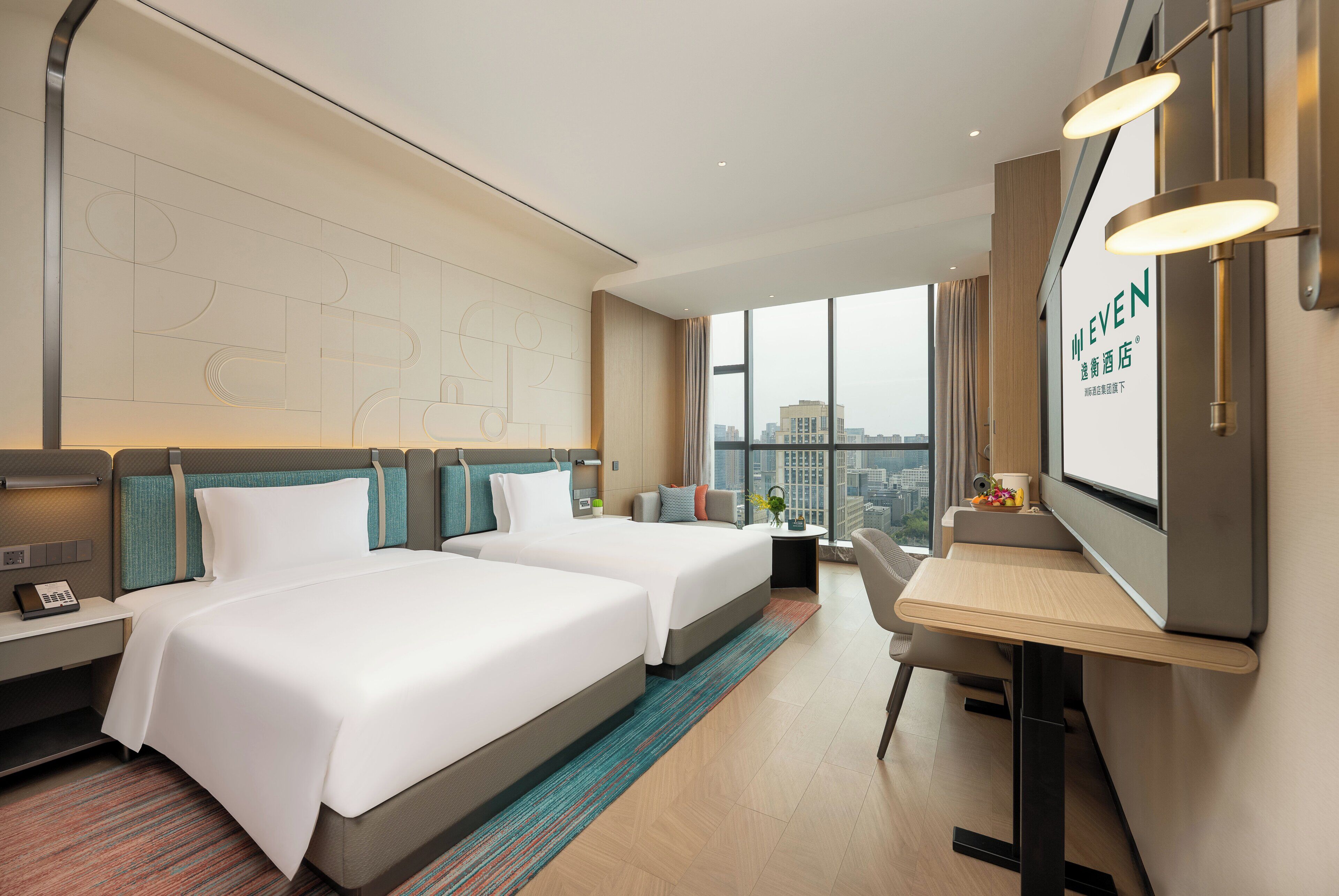 A hotel room with two beds, a desk, a chair, a TV, and a large window with a city view.