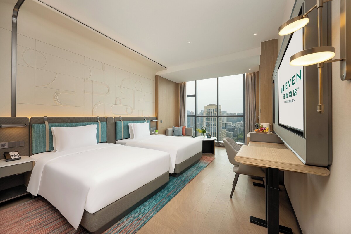 Hotelkamer met twee bedden, een bureau, een stoel, een televisie en een groot raam met uitzicht op de stad.