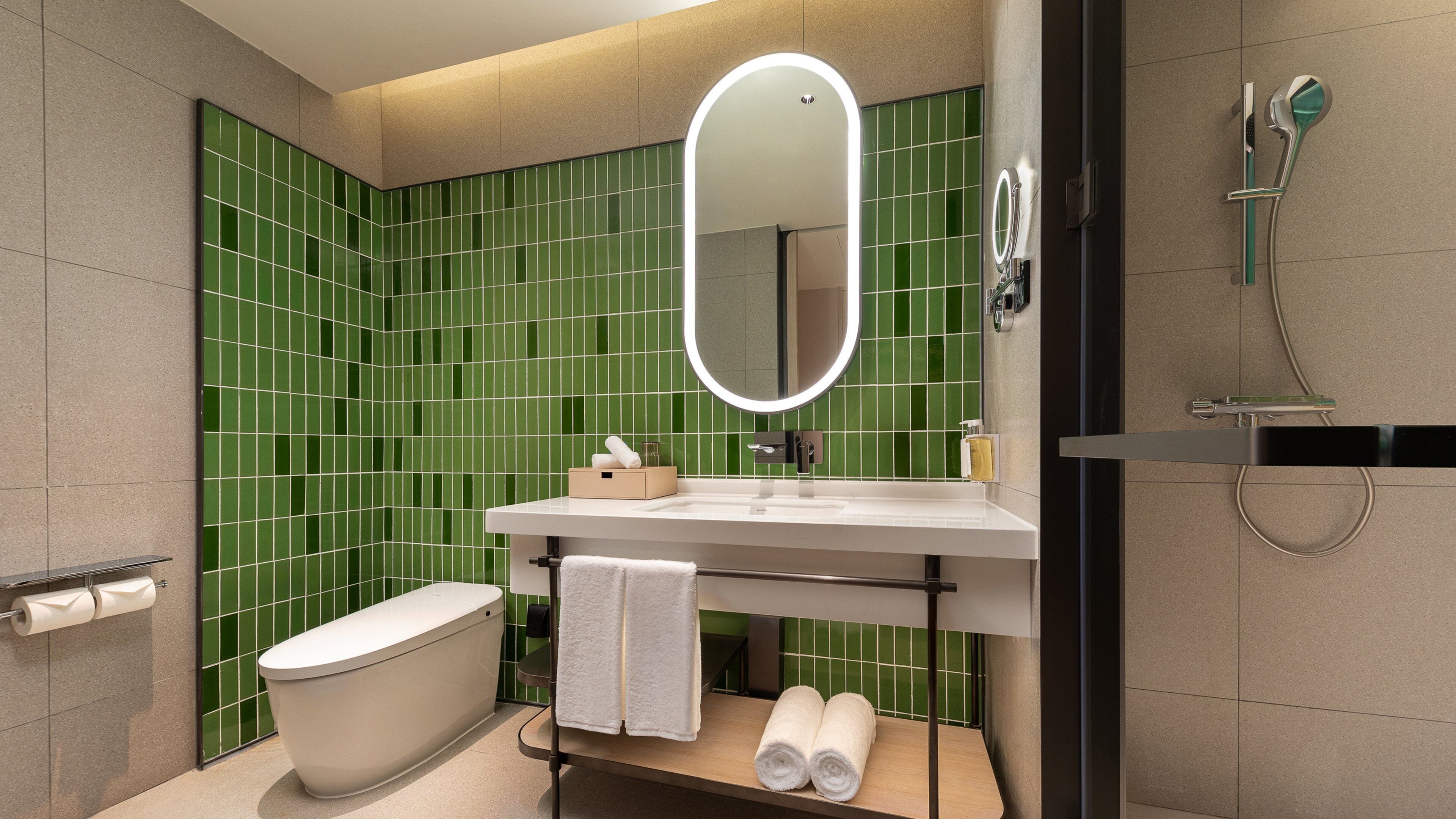 Un baño moderno con pared de azulejos verdes, un lavamanos blanco, un espejo, un inodoro y una zona de ducha.