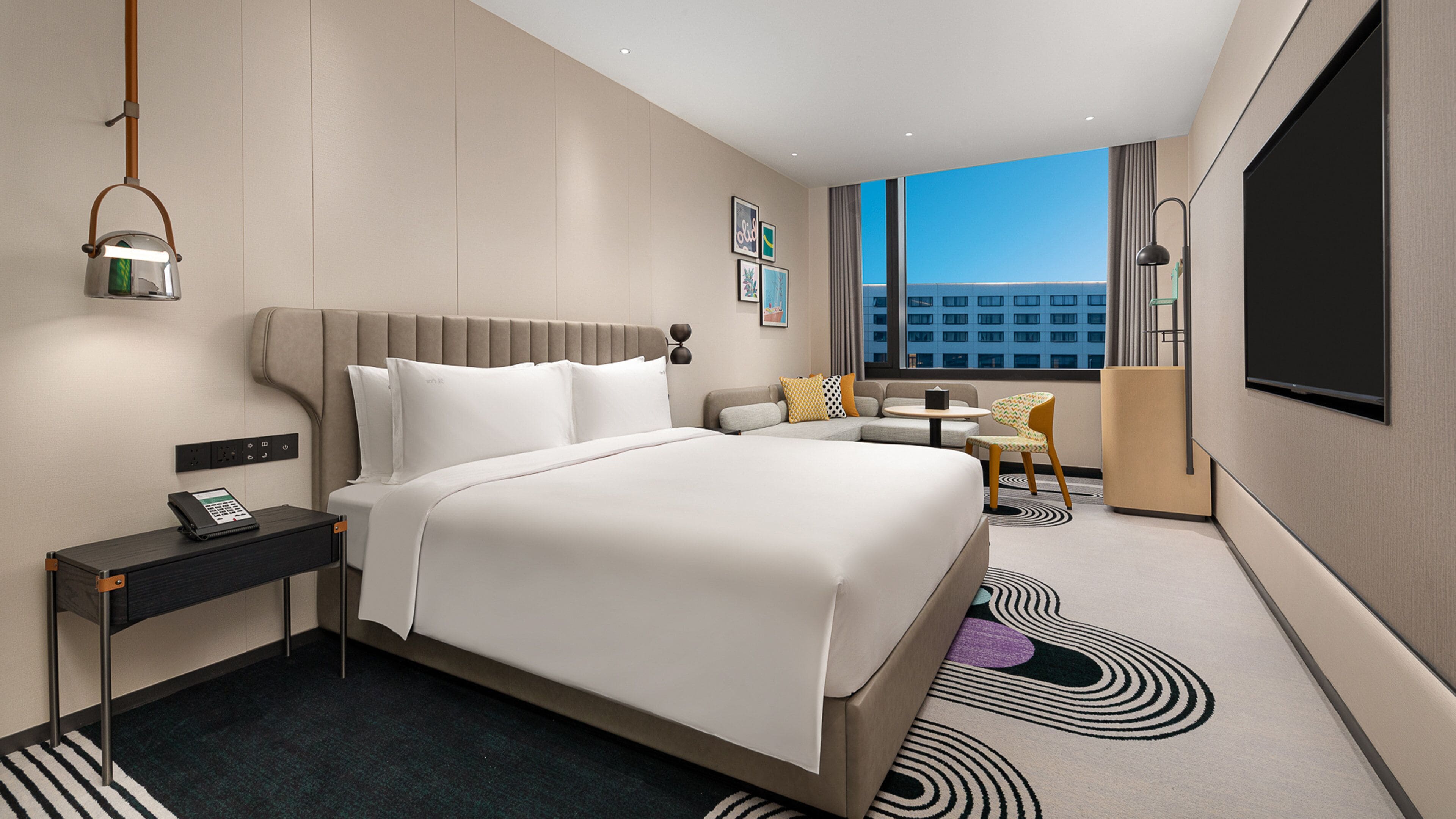 Habitación de hotel moderna con una cama grande, mesita de noche con teléfono, una pequeña mesa redonda con una silla y vistas a la ciudad a través de amplias ventanas.