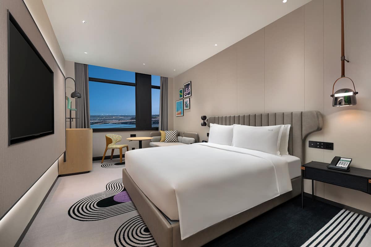 A modern hotel room with a large bed, a desk, a chair, and a view of the cityscape through the window.