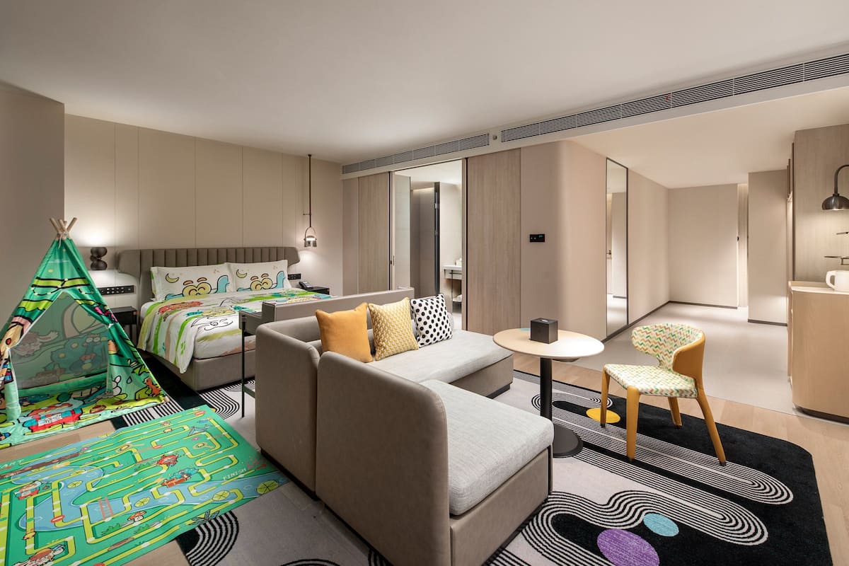 A hotel room with a bed, a sofa, a small table, a chair, a kitchenette, and a bathroom.