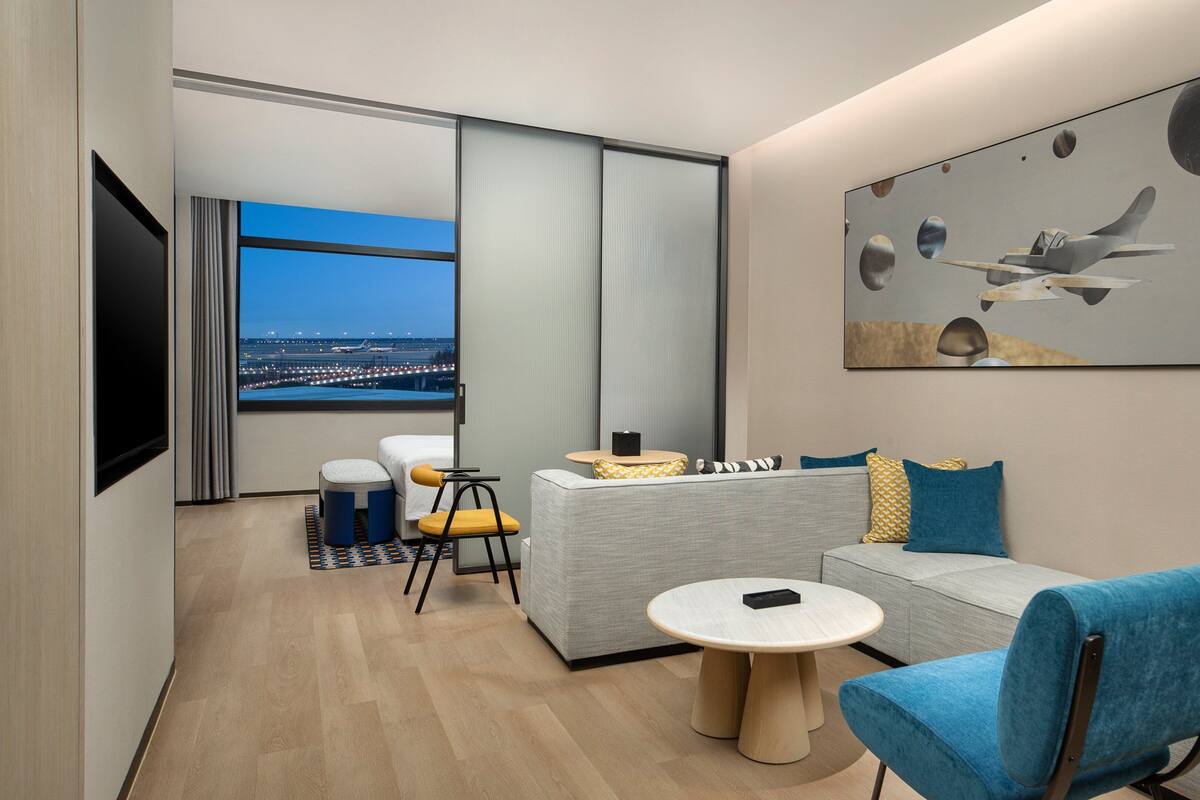 A modern hotel room with a sofa, two armchairs, a small round table, and a large window offering a city view.