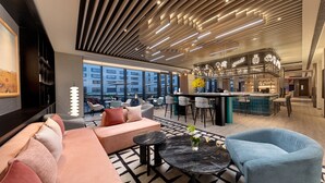 Un vestíbulo moderno de hotel con un sofá, sillas, una zona de bar y ventanas amplias que ofrecen vistas de la ciudad.