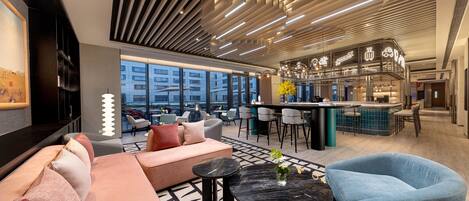 A modern hotel lobby with a sofa, chairs, a bar area, and large windows offering a view of the city.