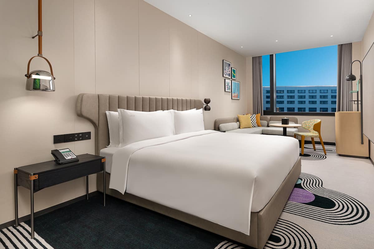 A modern hotel room with a large bed, bedside table with a phone, a small round table with a chair, and a view of the city through large windows.
